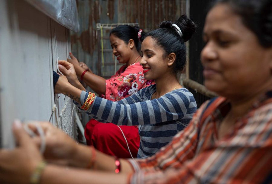 Drei Frauen, die an einer Wand Handarbeiten ausführen, vielleicht Stickerei oder Näharbeiten, mit einem Fokus auf die mittlere Person, die lächelt und deren Gesicht gut sichtbar ist. Sie tragen traditionelle und lässige Kleidung und arbeiten gemeinsam in einer gemütlichen, informellen Umgebung.