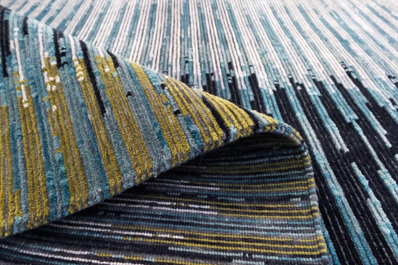 Texturierte Nahaufnahme eines gemusterten Teppichs mit vertikalen Streifen in verschiedenen Blautönen, Grau und Akzenten in Gelb.