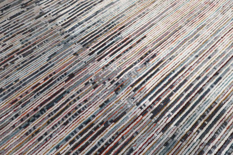 Texturierter Teppich mit einem vielfarbigen Streifenmuster, das sich in einer leichten Diagonale über das Bild zieht. Farben variieren von Rottönen über Blau und Grau bis hin zu Beige und Weiß, mit gelegentlichen dunklen Akzenten.