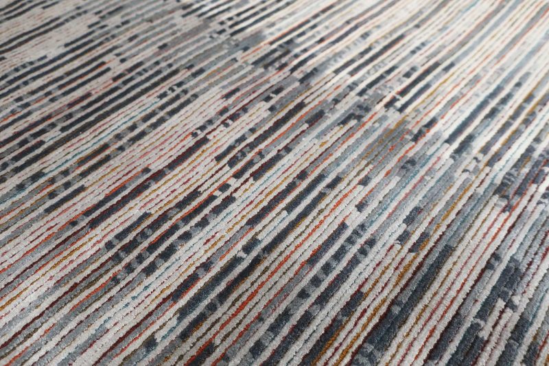 Texturierter Teppich mit einem vielfarbigen Streifenmuster, das sich in einer leichten Diagonale über das Bild zieht. Farben variieren von Rottönen über Blau und Grau bis hin zu Beige und Weiß, mit gelegentlichen dunklen Akzenten.