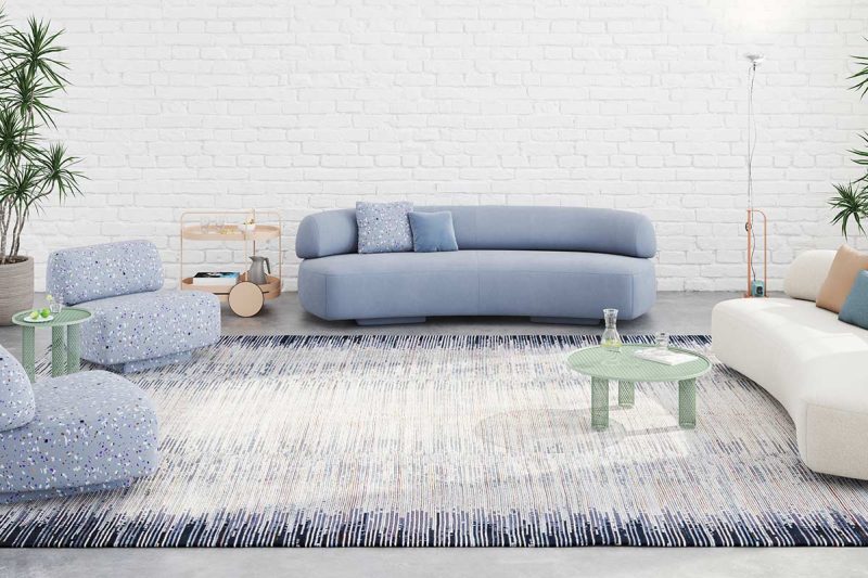 Modernes Wohnzimmer mit weißer Ziegelwand, großem Fenster mit Stadtblick, zwei großen Zimmerpflanzen in Töpfen, einem blauen Sofa, einem weiß-beigen Sessel, einem gemusterten Teppich und minimalistischen Möbeln wie einem Metallregal und runden Beistelltischen.