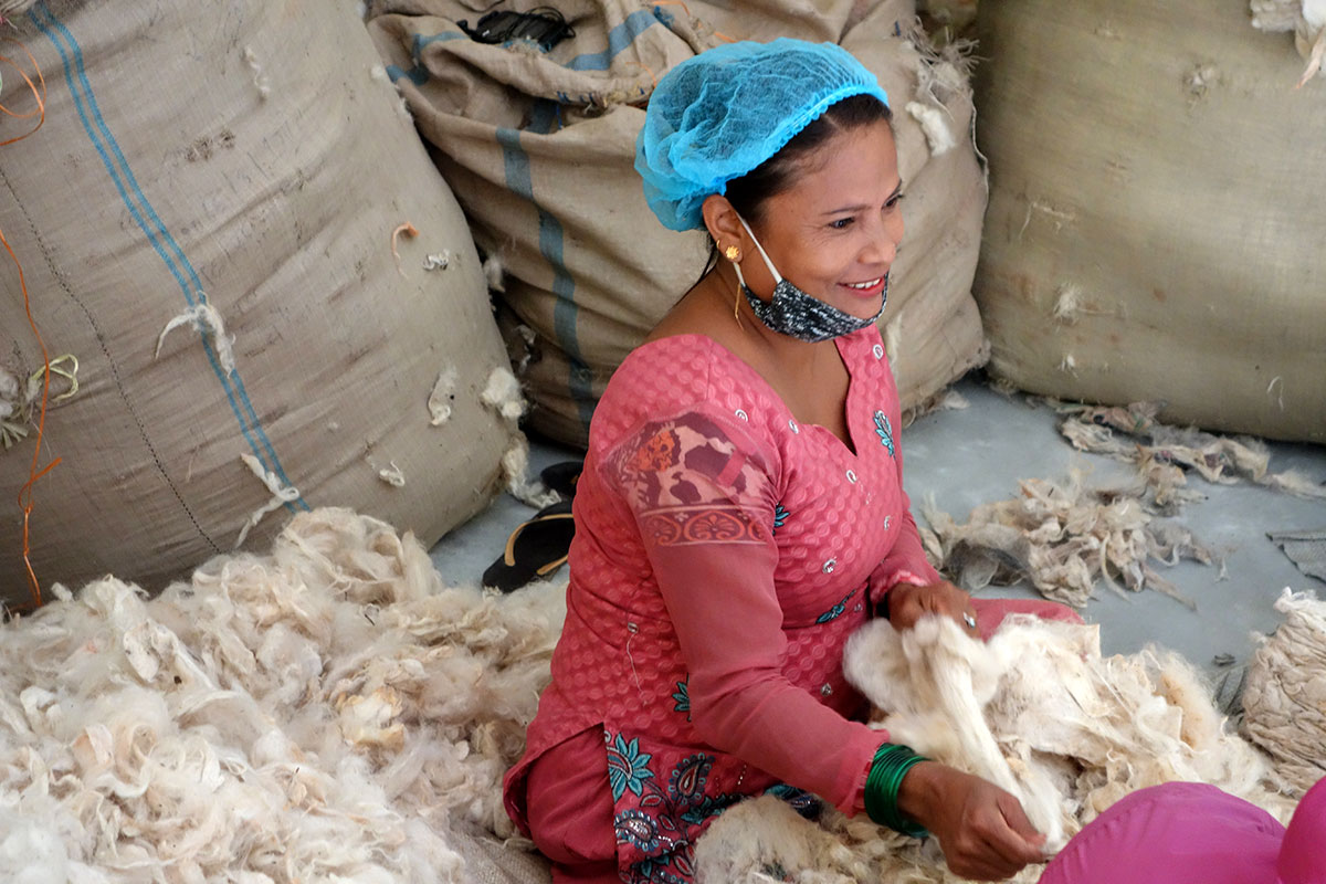 Frau in traditioneller Kleidung sitzt auf dem Boden und sortiert Rohwolle neben großen Säcken in einer Verarbeitungsanlage.