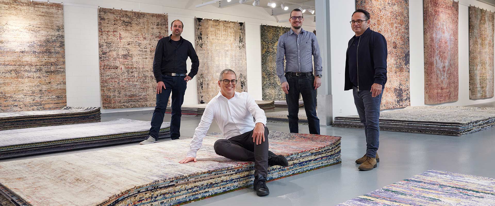 Vier Männer in einer Teppichgalerie mit mehreren aufgehängten und gestapelten Teppichen verschiedener Muster und Farben. Einer sitzt im Vordergrund auf einem Stapel Teppiche, drei stehen im Hintergrund und lächeln in die Kamera.
