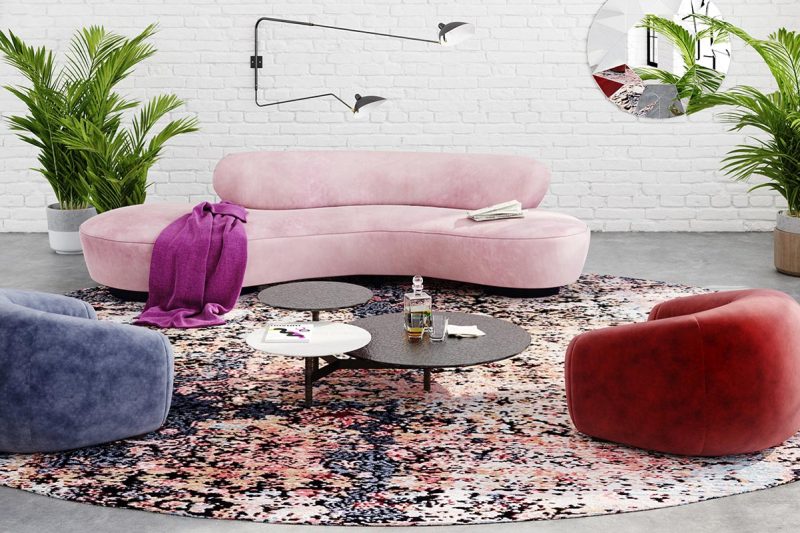 Modernes Wohnzimmer mit heller Ziegelmauer, großer geschwungener rosa Couch mit lila Decke, runden Couchtischen, buntem Teppich, blauen und roten Sesseln sowie Zimmerpflanzen und dekorativen Wandelementen.