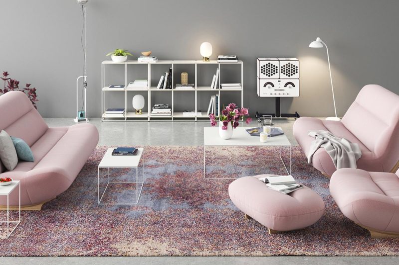 Modernes Wohnzimmer mit großer Fensterfront, heller Einrichtung, rosa Sesseln, weißem Bücherregal und Dekopflanzen, auf einem mehrfarbigen Teppich über hellem Boden.