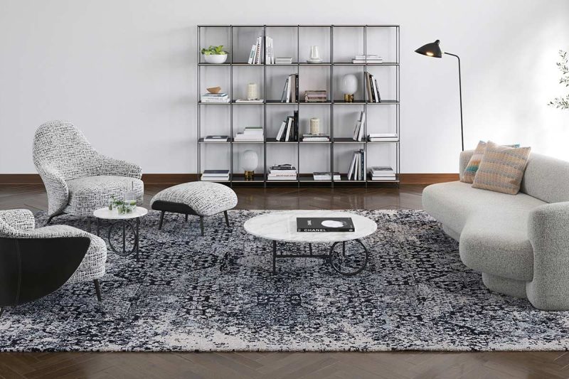 Modern eingerichtetes Wohnzimmer mit grauem Sofa, gemustertem Teppich, zwei weißen Couchtischen und Sesseln, Metallregal mit Büchern, Stehleuchte und Zimmerpflanze.