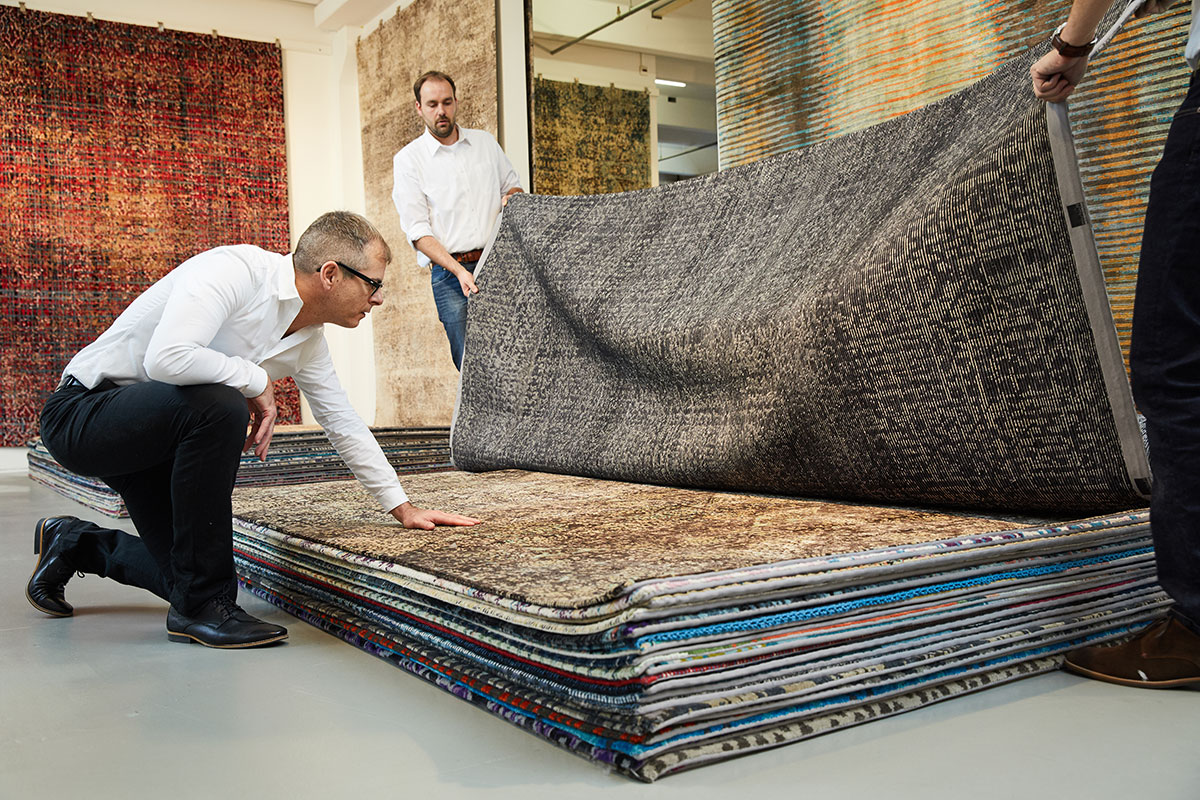 Drei Männer in einem Teppichgeschäft, einer kniet und streicht über einen Stapel Teppiche, während die anderen zwei einen Teppich hochheben, um die Muster zu präsentieren.