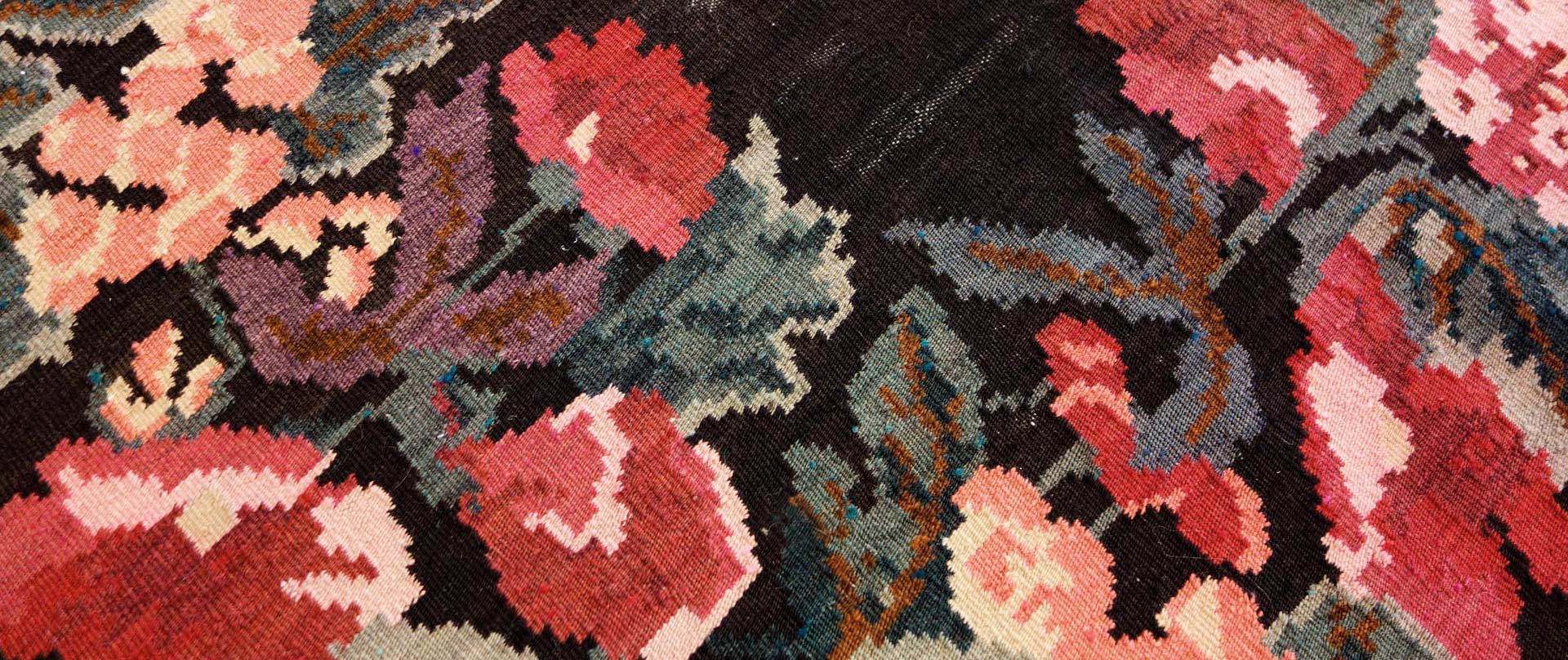 Detailaufnahme eines gemusterten Teppichs mit floralen Motiven in verschiedenen Farben, hauptsächlich in Tönen von Rosa, Blau und Schwarz.