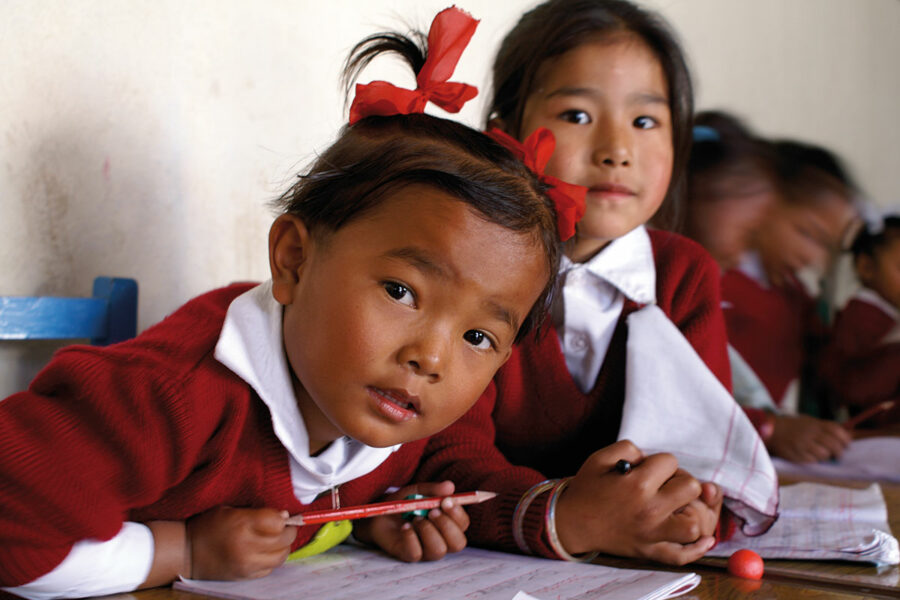 Zwei Kinder in Schuluniformen, ein Junge und ein Mädchen, sitzen in einem Klassenzimmer und arbeiten an ihren Aufgaben; der Junge blickt direkt in die Kamera, während das Mädchen lächelnd zur Seite schaut.
