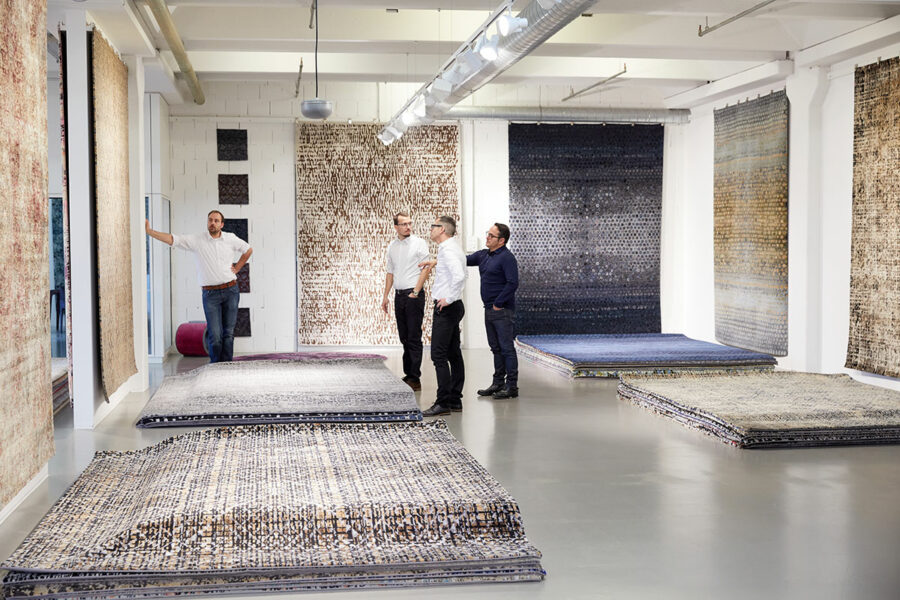 Innenraum eines Teppichgeschäfts mit mehreren ausgestellten Teppichen, die auf dem Boden und an den Wänden präsentiert werden; vier Männer betrachten und diskutieren über die Produkte.