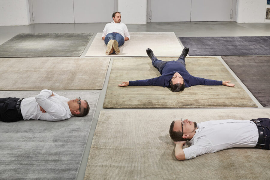 Vier Männer liegen entspannt auf dem Rücken verteilt auf verschiedenen Teppichen in einem großen, hellen Raum. Jeder Teppich hat ein unterschiedliches Design und Farbschema. Die Männer sind leger gekleidet, zwei in Hemden und Jeans, die anderen zwei in Hemden und Anzughosen.