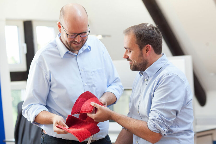 Zwei lächelnde Männer in bürotauglicher Kleidung betrachten und halten gemeinsam ein rotes Teepichmuster in einem hellen Raum.