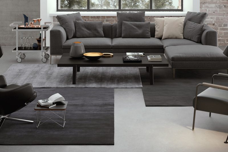Modern eingerichtetes Wohnzimmer mit grauem Ecksofa, dunklen Designerstühlen, einem Couchtisch und Beistelltischchen auf einem mehrschichtigen, grauen Teppich, belebt durch eine Ziegelwand im Hintergrund und metallische Deko-Elemente.