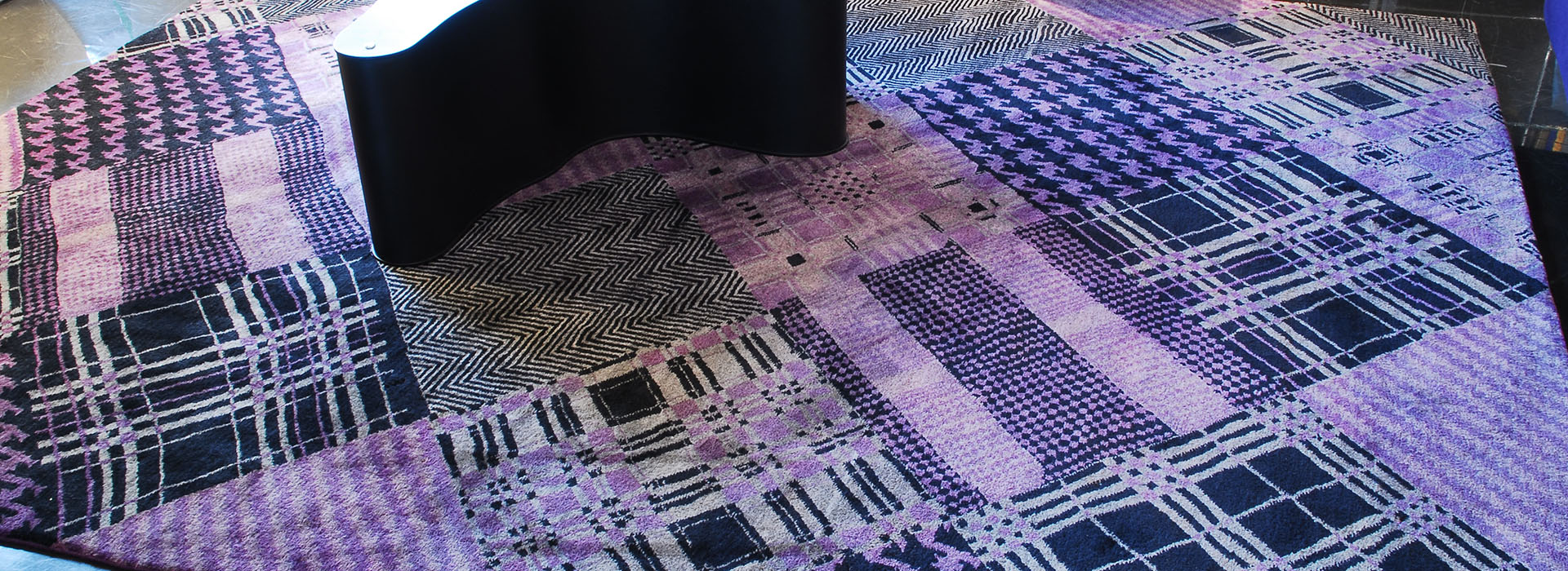 Texturierte Patchwork-Teppich mit abstrakten Mustern in verschiedenen Schattierungen von Lila, Schwarz und Weiß mit einer Ecke, die von einem dunklen Gegenstand überlappt wird.