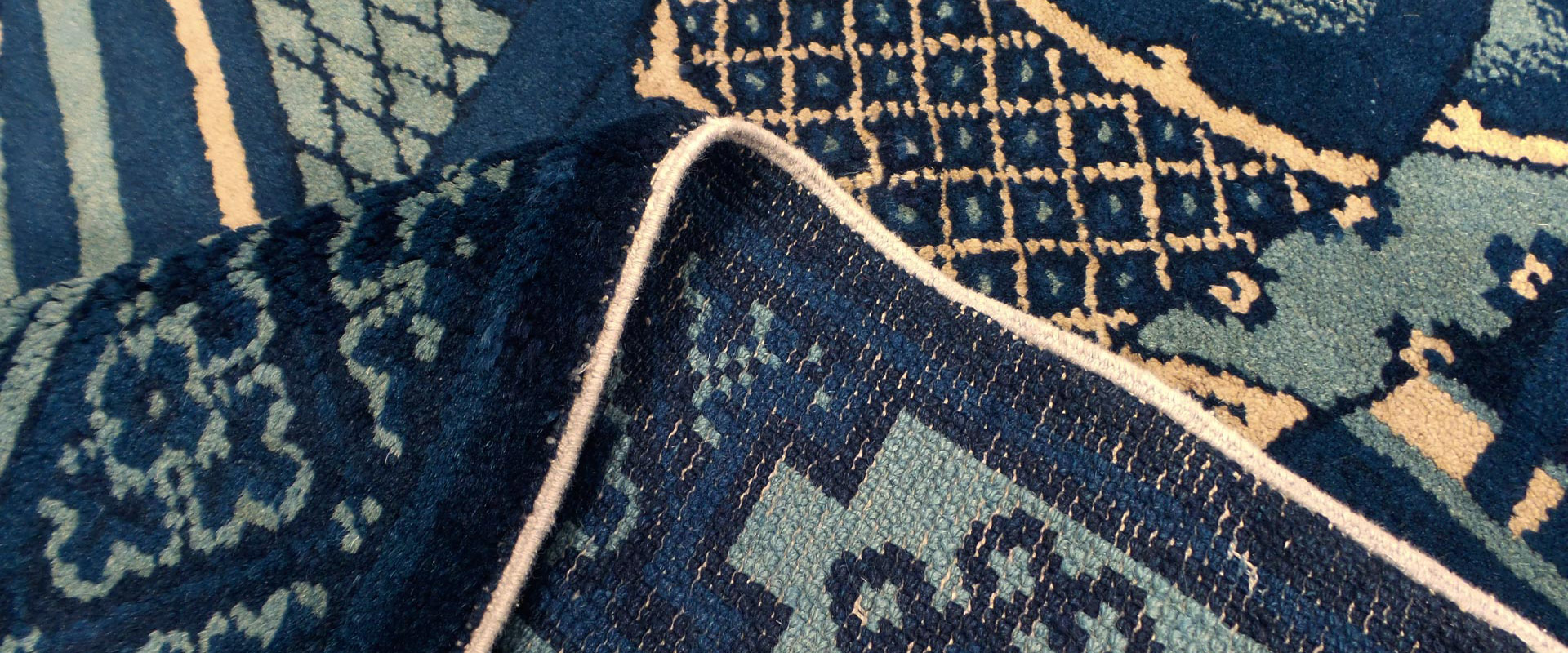 Nahaufnahme eines orientalischen Teppichs mit umgeschlagener Ecke, zeigt dunkelblaue und beige Muster auf türkisfarbenem Grund.