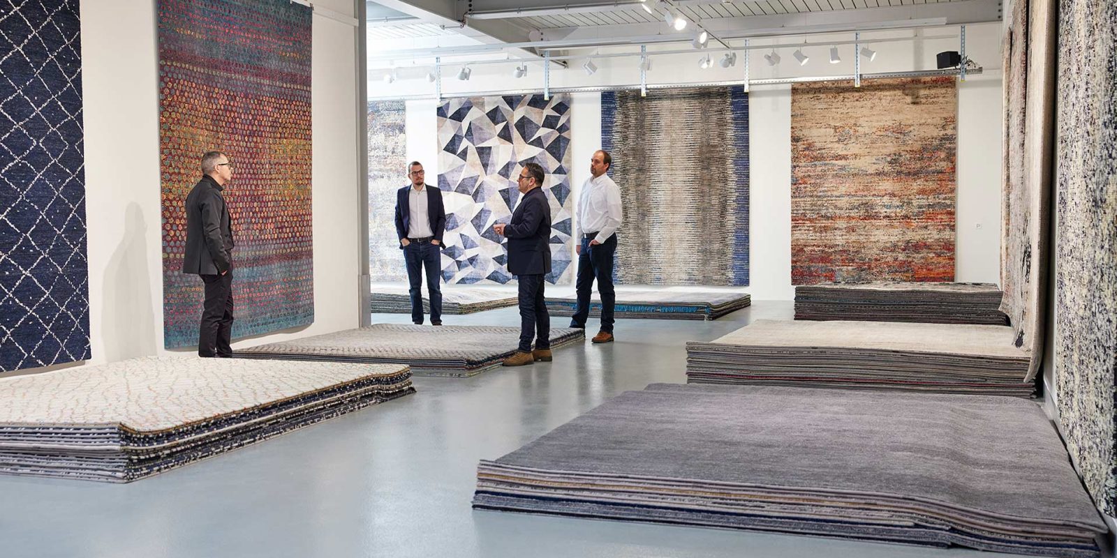 Vier Männer in einer modernen Teppichgalerie betrachten verschiedene hängende und gestapelte Designer-Teppiche.