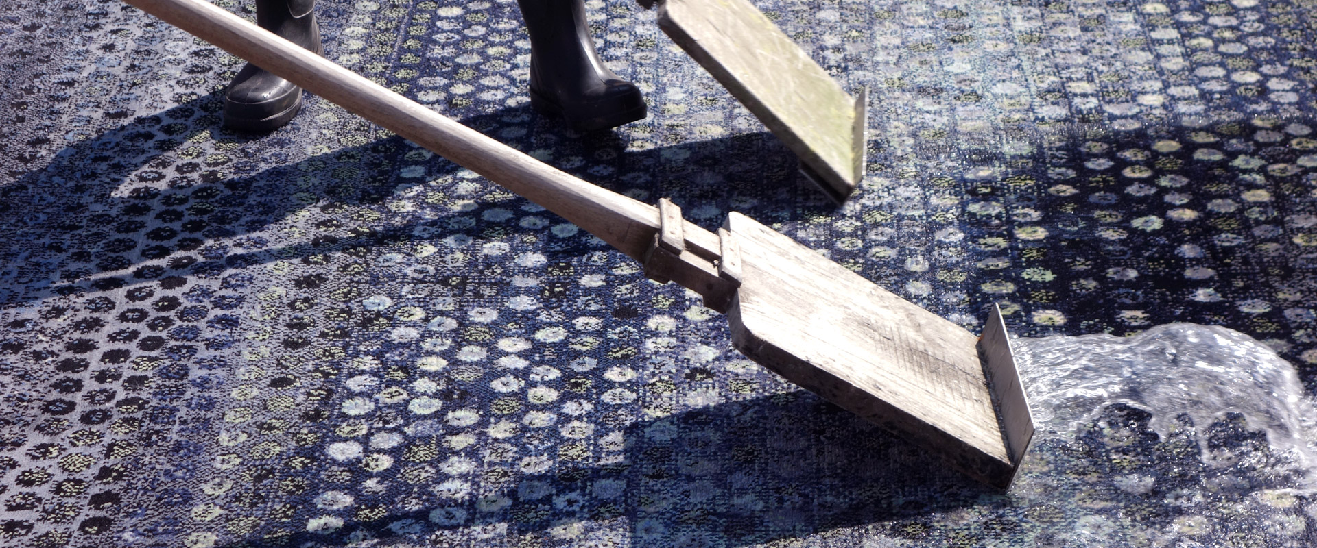 Nahaufnahme eines traditionellen Holzschaukelstuhls mit geschwungenen Kufen auf einem gemusterten Teppich im Sonnenlicht, wobei nur der untere Teil des Stuhls und der Füße einer Person sichtbar ist.