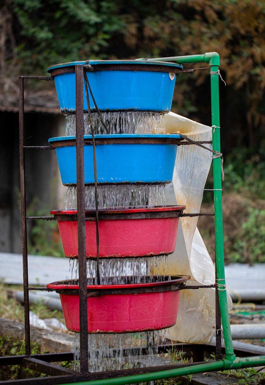 Eine gestapelte Anordnung von blauen und roten Plastikeimern, durch die Wasser fließt, aufgebaut auf einem metallischen Gestell vor einem natürlichen, grünen Hintergrund.
