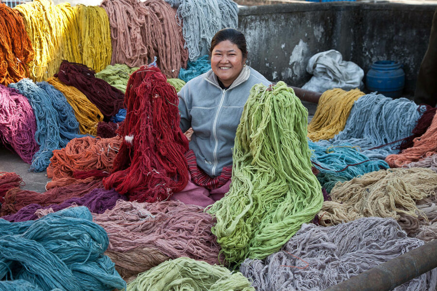 Frau sitzt lächelnd umgeben von zahlreichen Strängen bunt gefärbter Wolle, die zum Trocknen ausgelegt sind.