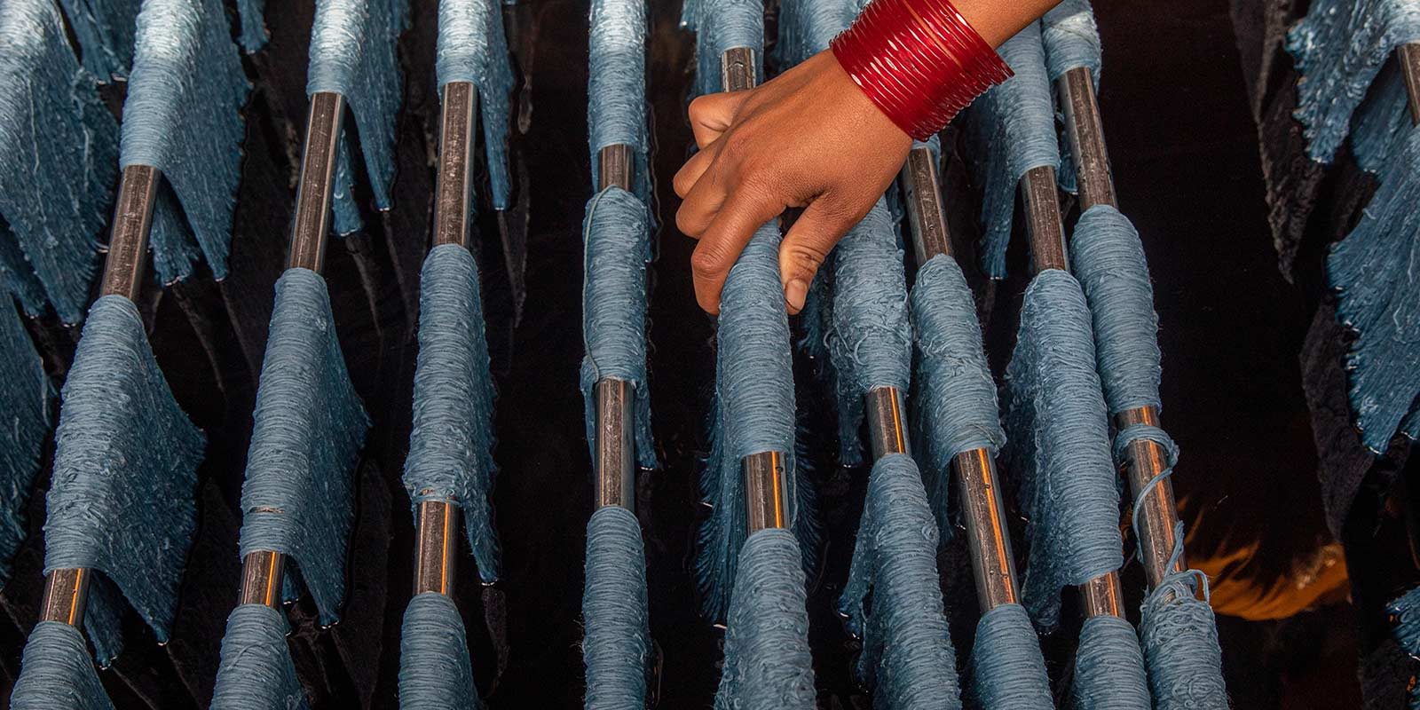 Hand mit rotem Armreif berührt blaue Garnrollen auf Metallstangen in einer Textilmanufaktur.